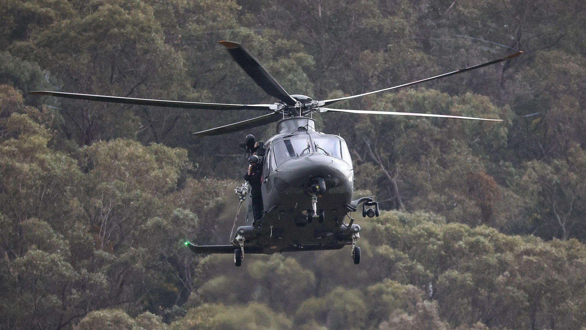 A police AW139 low over the tree canopy, a flight officer at the open door.
