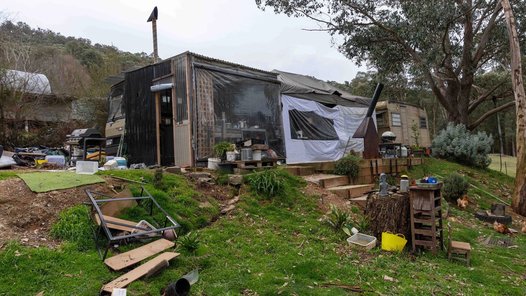 The bus and its corrugated annexe on the terraced hillside, tarps and camp life around it, chickens by the steps.
