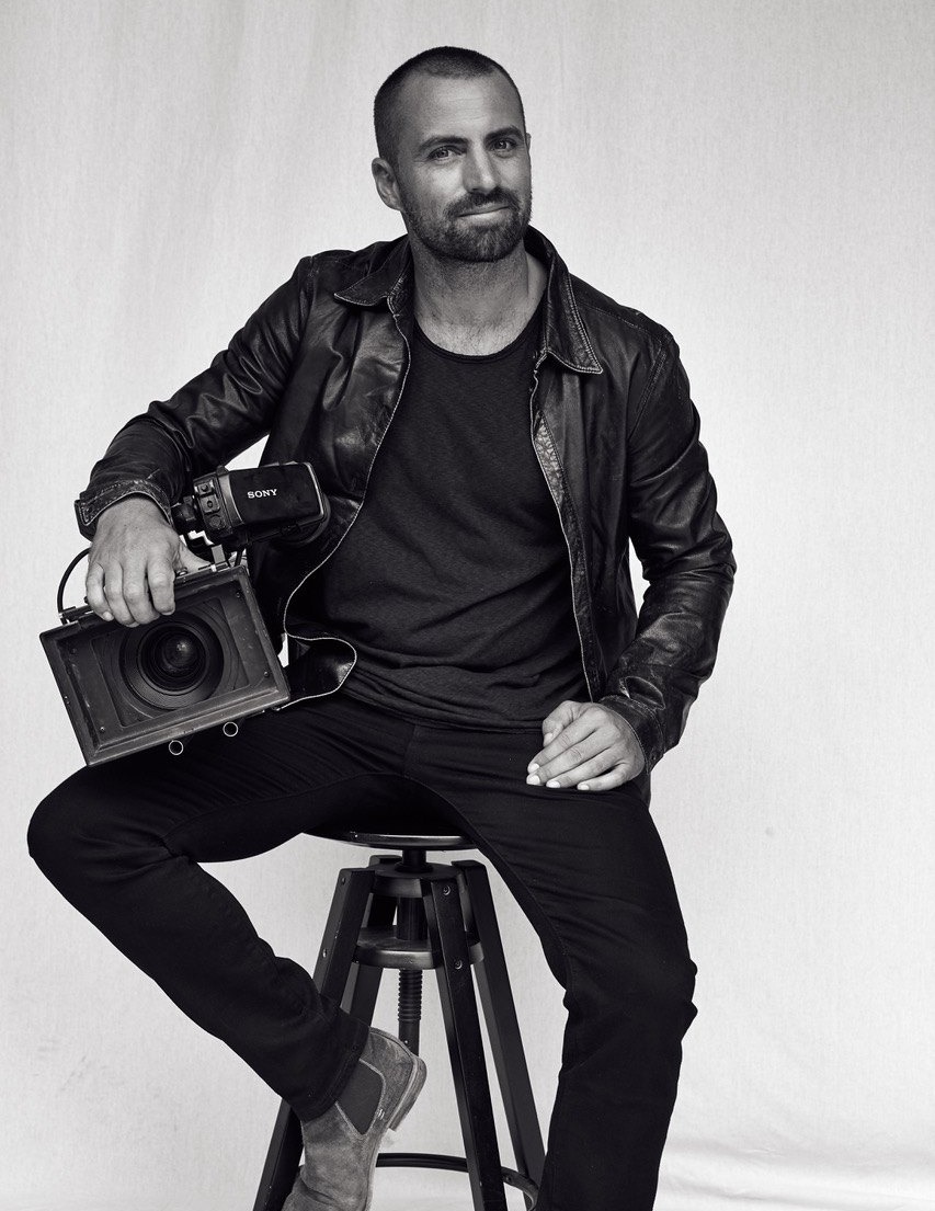 Tim Noonan seated with a cinema camera, black and white studio portrait.