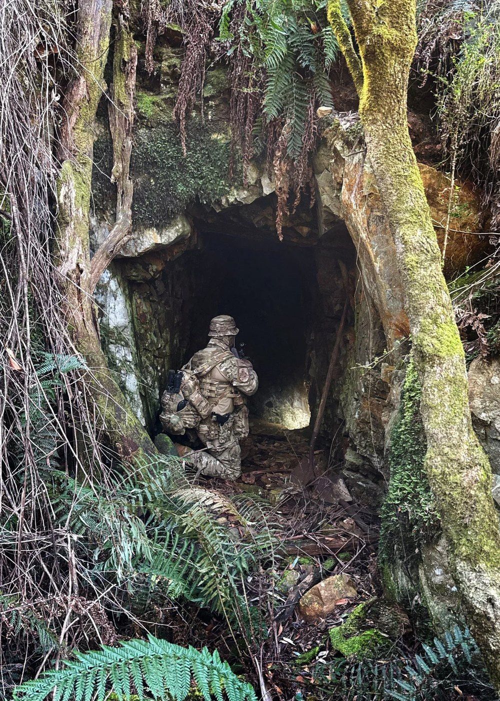 An officer kneeling at the fern-hung entrance of an old goldmine, weapon raised toward the dark.