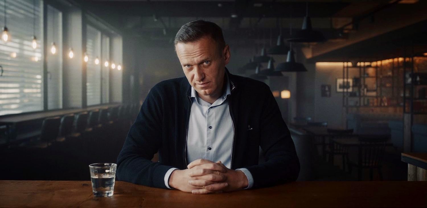Reference frame for interview camera A: a centred portrait, hands folded, the room part of the character.