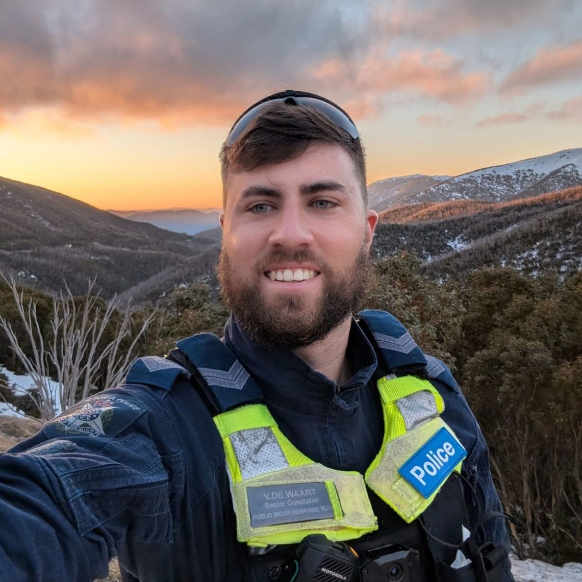 Vadim De Waart-Hottart smiling in police uniform on an alpine ridge at sunset, snow on the ranges behind him.